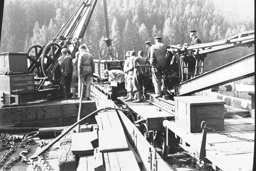 Das historische Schwarz-Weiß-Foto zeigt eine Gruppe von Arbeitern und Ingenieuren, die mit einem großen, Kran auf einer Eisenbahnbaustelle arbeiten. Die Szene spielt sich in einer waldreichen, bergigen Umgebung ab, während der Sanierungsarbeiten am Adlitzgraben Viadukt der Semmeringbahn.