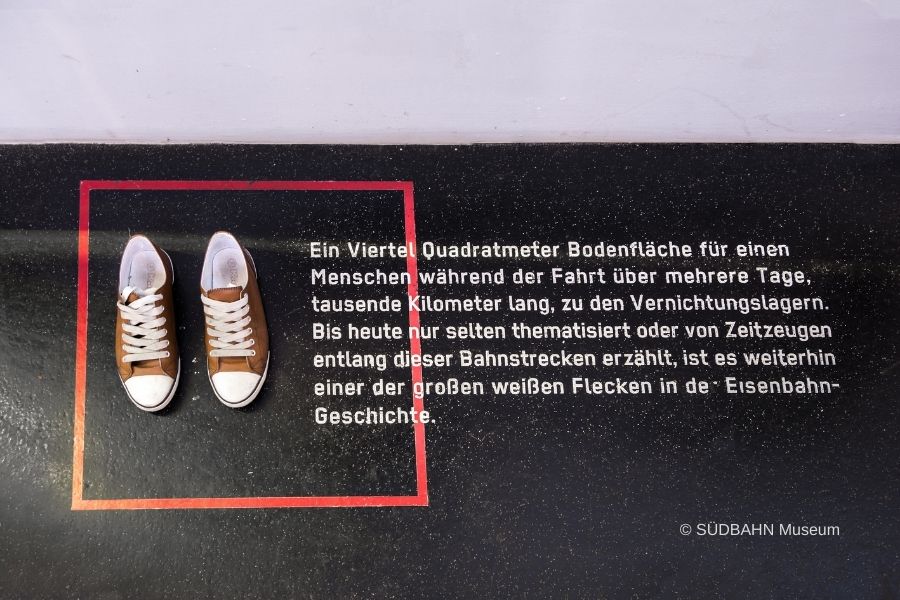 Das Foto zeigt den Ausstellungsbereich Zwischen den Fronten im SÜDBAHN Museum. 2 Mädchenschuhe stehen in einem roten Quadrat. Daneben ein Ausstellungstext.