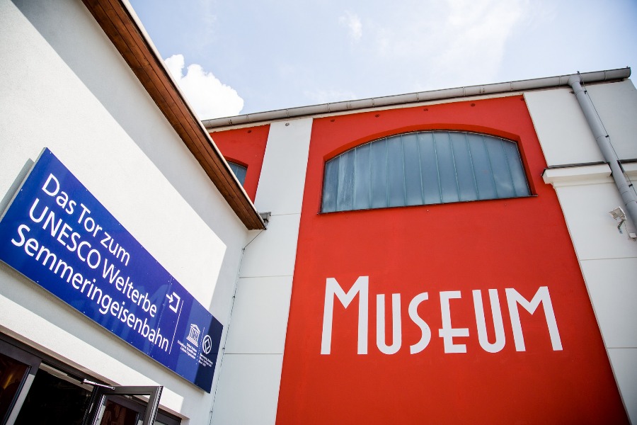 Das aktuelle Foto zeigt einen Teil der Eingangssituation des SÜDBAHN Museums. Eine blaue Tafel über der Eingangstüre und seitlich die rote Fassade des Gebäudes.