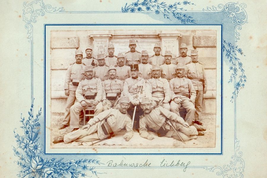 Das historische Foto zeigt eine Gruppe von Eisenbahnern in Uniform, die für ein Gruppenporträt vor dem Ghega Denkmal posieren, wobei einige sitzen, stehen oder liegen. Unten handschriftlich „Bahnwache Eichberg“ notiert.
