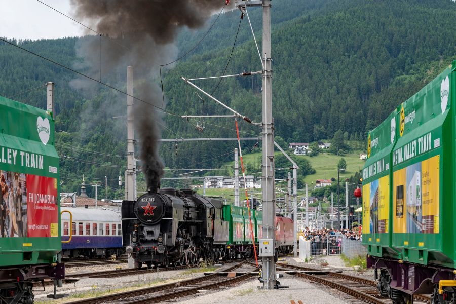 Das Foto zeigt einen Ausschnitt des Bahnhofes Mürzzuschlag. Rechts und links des Fotos sind grüne Waggons der Firma Innofreight zu erkennen. In der Bildmitte eine rauchende Dampflokomotive.
